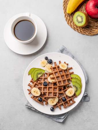 A freshly prepared vegan waffles with kiwi, blueberries and banana slices on a white plate with a cup of coffee and fresh fruits on a light background. Healthy dietary and delicious breakfast.の写真素材