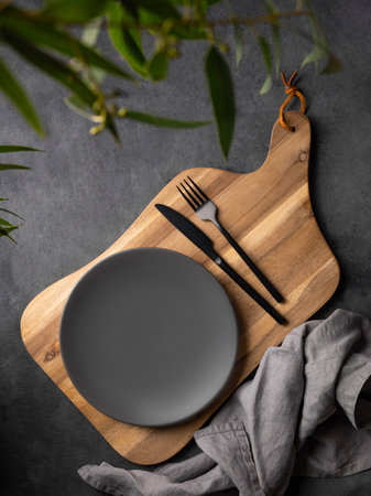 Empty gray plate on a wooden board with cutlery on a dark background with green leaves and a napkin. Modern showcase rustic concept with stage for product presentation, sale or food advertising bannerの写真素材