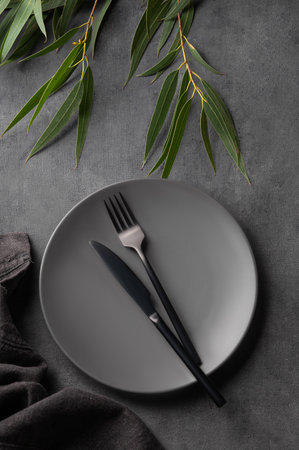 Festive table setting with empty gray plate and black cutlery for holiday with eucalyptus branches on dark background. Serving concept for restaurant menu. Top view.の写真素材