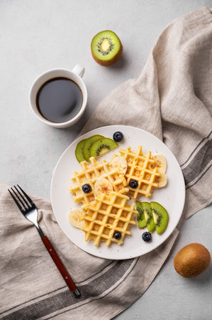 A freshly prepared waffles with kiwi, blueberries and banana slices on a white plate with a cup of coffee and a napkin on a light gray background. The concept of a healthy dietary and breakfast.の写真素材