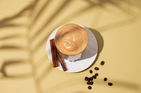 Dalgona coffee. Whipped instant coffee in a glass on a saucer with cinnamon sticks on yellow background with shadow leaf.  The concept of a trendy and popular drink.Top view.の写真素材