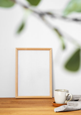 A  frame with a blank canvas against a light wall and on a wooden tabletop with blurred green foliage and cup of tea. Layout concept for photography, poster or painting. Copy space.の写真素材