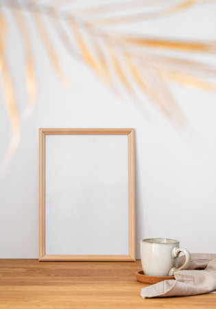 A frame with a blank canvas against a light wall and on a wooden tabletop with blurred palm leaf and cup of tea. Layout concept for photography, poster or painting. Copy space.の写真素材