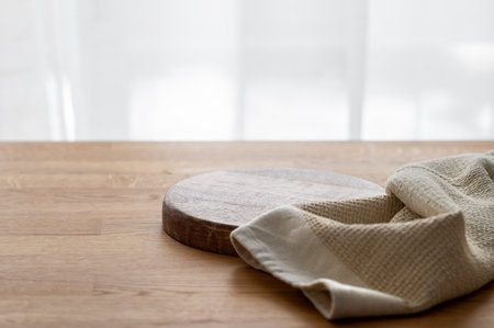 Empty cutting board on wooden texture tabletop with kitchen towel in front of window. Board for serving food or preparing homemade and healthy food. Front view and copy space.の写真素材