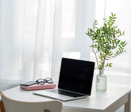 Desktop with laptop, notepad, phone and glasses in front of a window with morning sunlight. Home cozy office concept in Scandinavian style.の写真素材