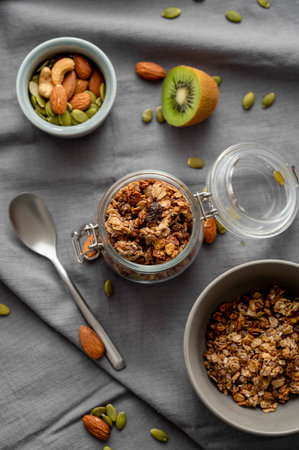Homemade granola in a jar and bowl on a dark linen background with nuts and fruits. The concept of healthy and nutritious whole grain muesli for breakfast. Top view.の写真素材