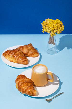 Coffee cup and fresh croissants on blue background with yellow flowers and shadow. Creative layout and concept of healthy food and french breakfast. Copy space.の写真素材