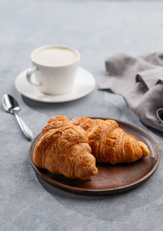 A cup of aromatic cappuccino coffee and fresh croissants in a wooden plate on a blue background with morning light. The concept of a delicious and healthy breakfast with homemade cakes. Copy space.の写真素材