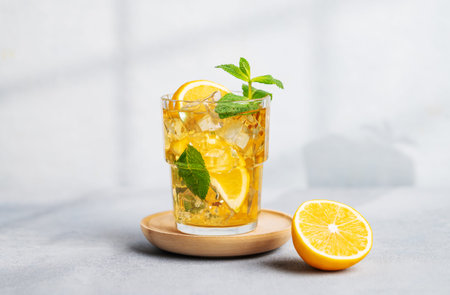 Iced tea with lemon, mint in a glass on light background with shadow and citrus fruits. The concept of a refreshing drink or lemonade on a hot summer day. Front view.