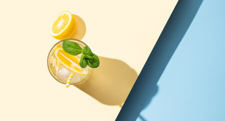 A  cold lemonade in a glass with lemon, mint and ice on a yellow and blue background with shadow. The concept of a refreshing and healthy drink.  Top view and copy space.の写真素材