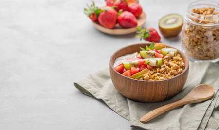 Natural yogurt with granola, kiwi and strawberries in a wooden bowl on a light background with fresh berries, fruits and napkin. Healthy and nutritious breakfast concept. Copy space.の写真素材