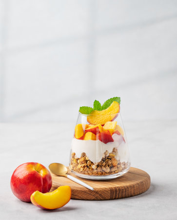 Yogurt parfait with peach and muesli in a glass on a wooden board on a light background with fresh fruits and shadow. Healthy eating concept. Nutritious summer breakfast. Copy space.の写真素材
