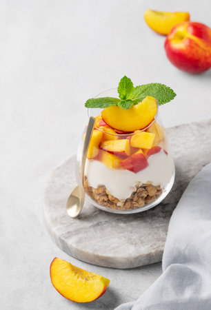 Greek yogurt parfait with peach and muesli in a glass on a marble board on a light background with fresh fruits and napkin. Healthy eating concept. Nutritious summer breakfast. Copy space.の写真素材