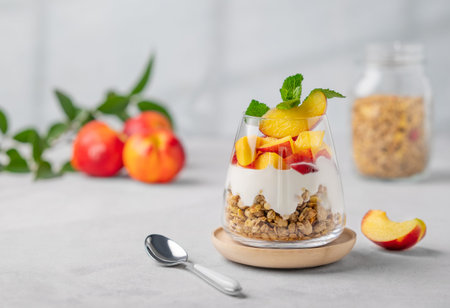 Greek yogurt parfait with peach and granola in a glass on a light background with fresh fruits and spoon. Healthy eating concept. Nutritious summer breakfast. Copy space.の写真素材
