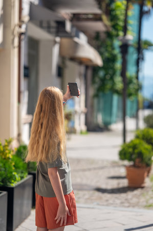 Young girl with long blonde hair taking selfie on street in city. Tourist standing and taking photo during summer vacation on blurred background in natural sunlightの写真素材