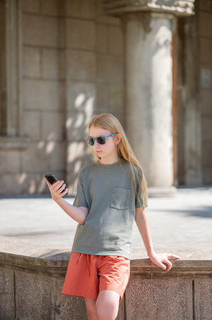 A young girl with long blond hair, wearing sunglasses, a green T-shirt and shorts, stands on the street in front of a historic building and holds a smartphoneの写真素材