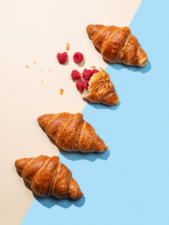 Flat lay of croissants with fresh raspberries on yellow and blue background with shadow. Creative...