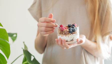 Granola with greek yogurt, raspberries and blueberries in a glass in hands of a woman with blond...