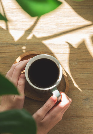 Woman hand holding mug with espresso coffee on a wooden background with  monstera leaves and shadows in bright sunlight. Morning energy drink concept. Top view and copy space.の写真素材