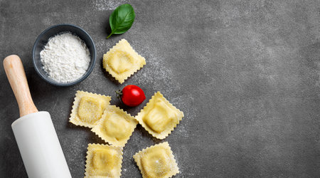 Flat lay of pasta ravioli with tomato and basil on dark background with flour and rolling pin. Traditional homemade italian food concept. Top view and copy space.の写真素材