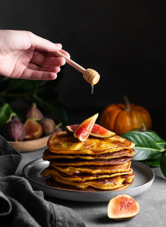 Hand holding honey stick and pouring it on stack of pumpkin pancakes with figs on dark background with fresh fruits. Concept of cozy home autumn food. Copy space.の写真素材
