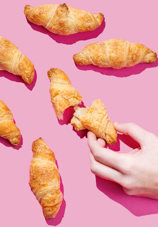 Hand holding fresh torn mini croissant on pink background with shadow. Design flat lay concept for breakfast pastry mockup or banner. Top view.の写真素材