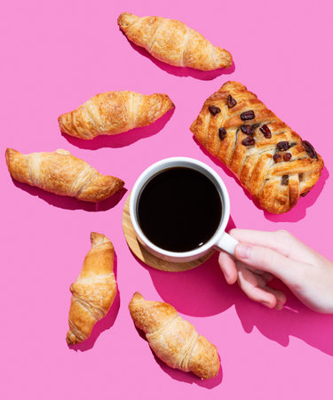 Hand holding a cup of espresso coffee on a pink background with fresh mini croissants and maple pecan bun. Design concept for a breakfast pastry mockup or banner. Top view.の写真素材