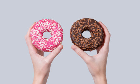 Hands holding two glazed chocolate and pink donuts on blue background. Creative concept of mockup or advertisement of fast breakfast. Copy space.の写真素材