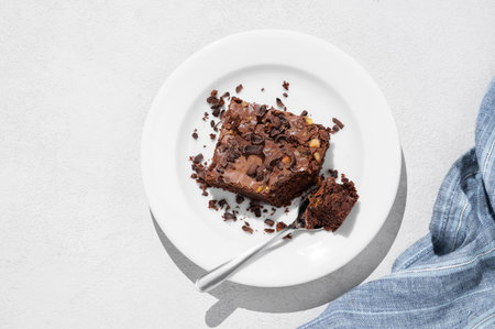 Chocolate brownie with nuts on a plate with spoon on a light background with morning shadow and napkin. Homemade sweet baking concept. Top view and copy space.の写真素材