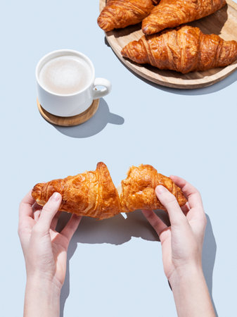 Hands breaking a fresh croissant over a blue background with a cup of cappuccino or latte coffee and pastry. Creative mockup for cafe advertising or French breakfast concept. Top view and copy space.の写真素材