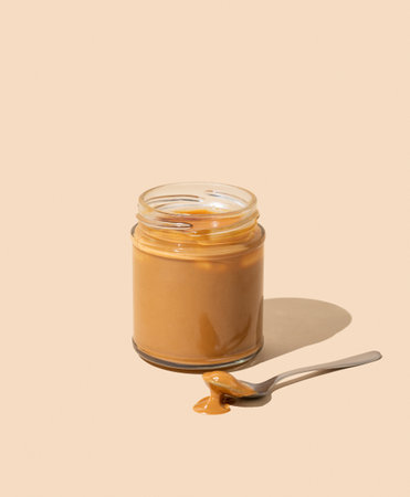 Peanut butter or caramel sauce in glass jar on isolated beige background with spoon and shadow. Concept of tasty and healthy snack for breakfast. Free space for text.の写真素材