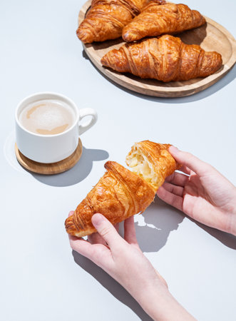 Hands breaking a fresh croissant on  a blue background with a cup of cappuccino or latte coffee and pastry. Creative mockup for cafe advertising or French breakfast concept.の写真素材