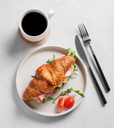 Croissant sandwich with ham, cheese, tomato  and arugula on a plate on a light background with a cup of  coffee. Concept for a healthy breakfast or brunch cafe menu. Top view.の写真素材
