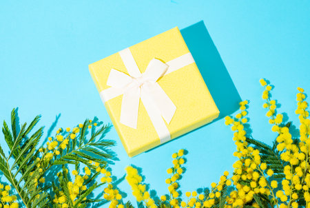 Yellow gift box on blue background with mimosa flowers and sunlight. Concept of advertising banner or greeting card for Mother's Day or spring holidays. Top view and copy space.の写真素材