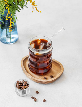 Cold coffee in a glass with ice on a wooden plate on a light background with a bouquet of spring mimosa and coffee beans. The concept of a craft refreshing and invigorating drink. Copy space.の写真素材