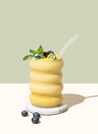 Smoothie with mango, banana and blueberry in a glass on a yellow and green background with berries and shadow.の写真素材