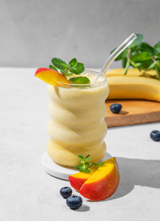 Milk smoothie with mango, banana and blueberry in a glass on a light background with tropical fruit slices and shadow. Healthy eating and detox diet concept. Front view and copy space.の写真素材