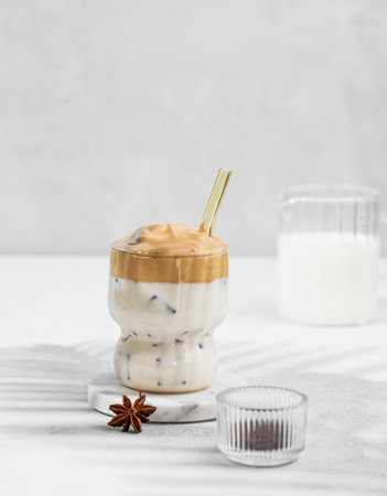 Dalgona coffee in a glass on a light background with milk jug, spices and shadow. Korean whipped drink with instant coffee, sugar, milk and ice. Sweet trendy dessert concept. Front view and copy spaceの写真素材