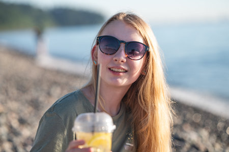 Smiling young woman with sunglasses holding a cold drink with lemon slices on a pebble beach, enjoying a sunny day by the seaの写真素材