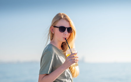 Young woman in sunglasses and casual t-shirt enjoying cold drink with lemon slices. Bright summer day on the beach, relaxation concept. Copy space.の写真素材