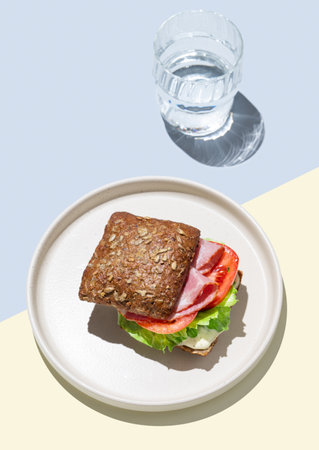 Toast sandwich made of grain bread with prosciutto, tomatoes, lettuce and sauce on blue yellow background with glass of water. Healthy breakfast concept for cafe menu advertising or banner. Top view.の写真素材
