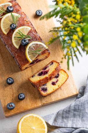 Lemon pie with sugar glaze, blueberries, decorated with rosemary and citrus slices on a wooden board on a light background. The concept of delicious homemade baking with fruits and berries. Top view.の写真素材