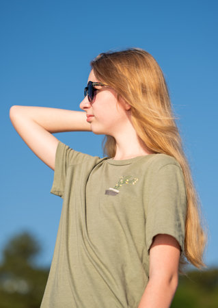 Young woman with long blonde hair wearing sunglasses and a green t-shirt, smiling under a clear blue sky. Capturing a happy and relaxed momentの写真素材