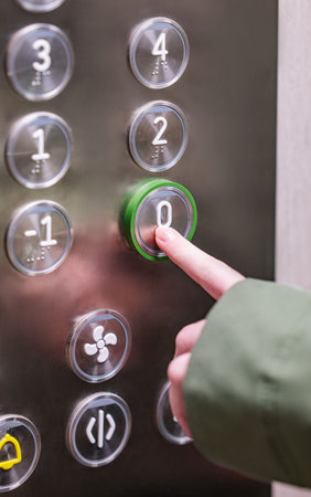 Close-up of hand pressing green-lit ground floor button in metallic elevator panel, showing floor...