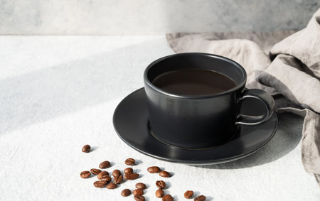 Black cup of  coffee espresso on light textured background with napkin, coffee beans and morning light from window. Aesthetic concept of breakfast drink. Front view and copy space.の写真素材