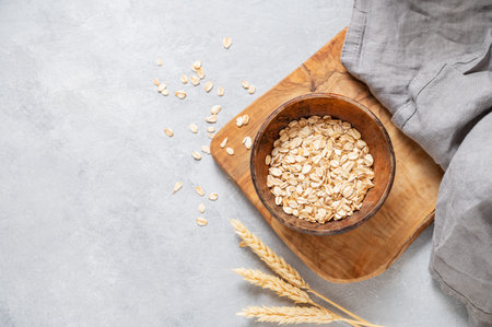 Oat flakes in bowl on wooden board with wheat and napkin on light textured background. Healthy...