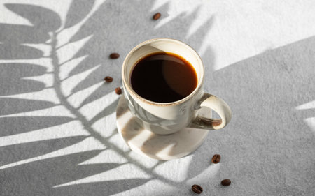 Cup of freshly brewed coffee on light textured background with shadow of palm leaf, coffee beans and morning light from window. Aesthetic concept of breakfast drink. Top view and copy space.の写真素材