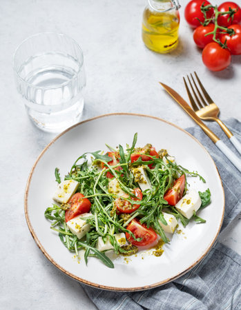 Healthy arugula salad with cherry tomatoes, mozzarella and pesto sauce on a plate on a light textured background with glass of water. Fresh and healthy Mediterranean food concept. Top view.の写真素材