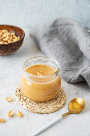 Glass jar with creamy peanut butter on grey background with scattered roasted nuts, spoon and...