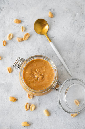 Glass jar  with creamy peanut butter with spoon on grey background with scattered roasted nuts and natural light. Natural homemade nut butter concept. Top view and copy space.の写真素材